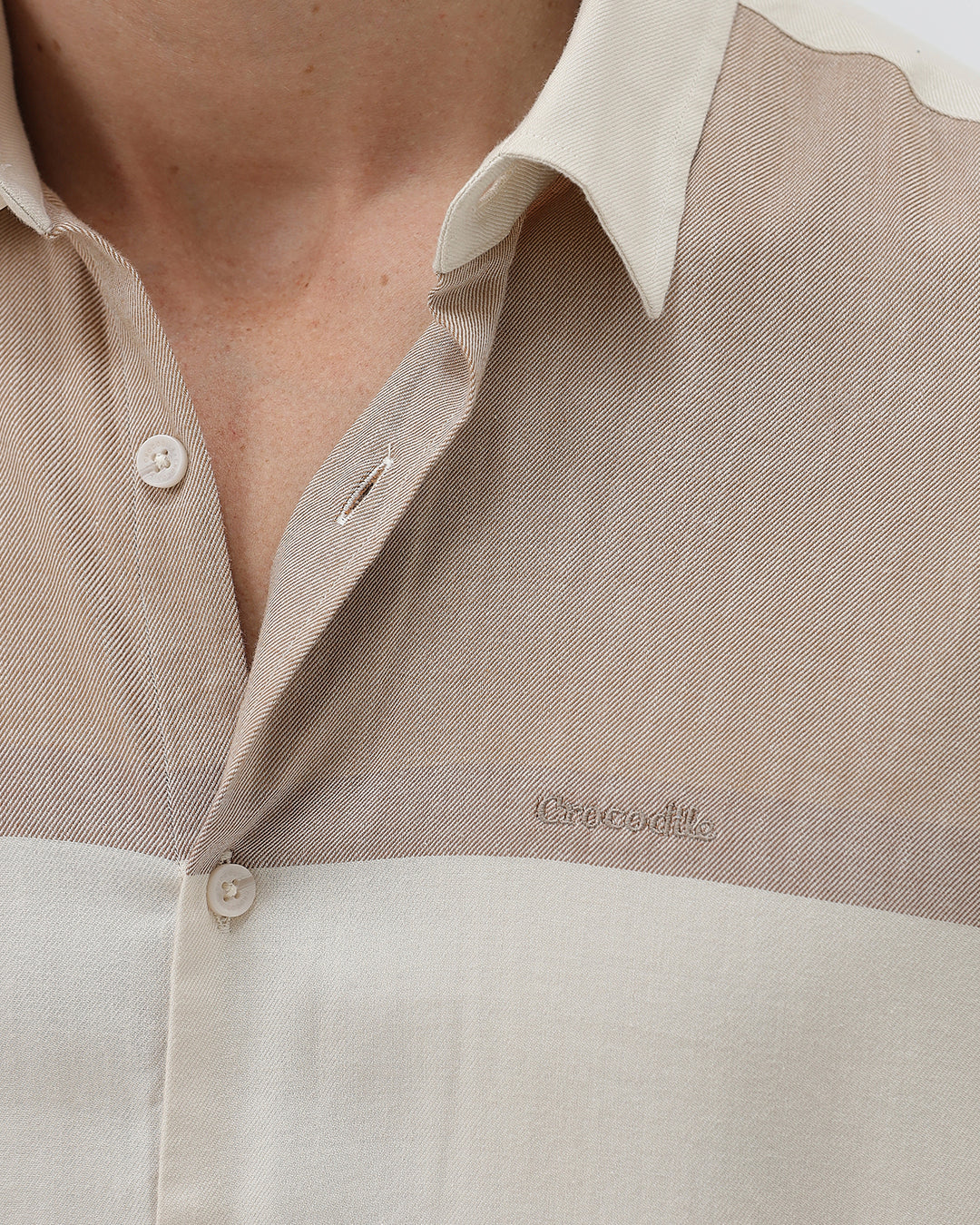 A man wearing a short-sleeve linen button-up shirt with a color-blocked design in beige and cream, paired with black pants, against a plain white studio background.