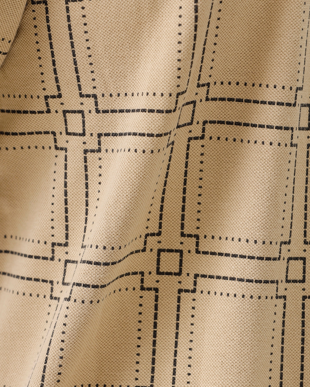A man wearing a beige short-sleeve polo shirt with a brown grid pattern, paired with black jeans, standing against a plain white studio background.