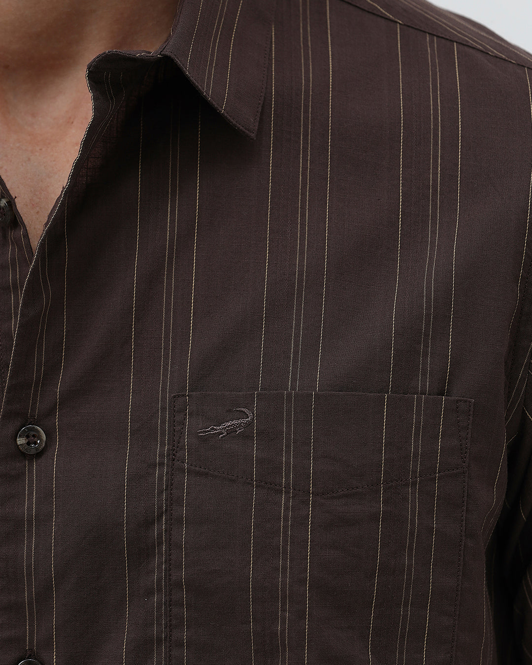 A man wearing a dark brown long-sleeve button-up shirt with thin vertical stripes and light gray trousers, standing against a plain light gray background.