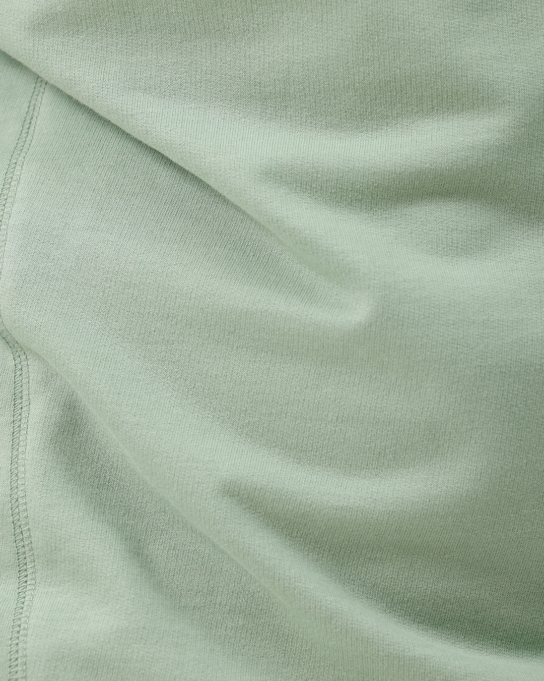 A man wearing a light sage green athletic t-shirt with a small tonal logo on the chest, paired with beige pants, standing against a plain white studio background.