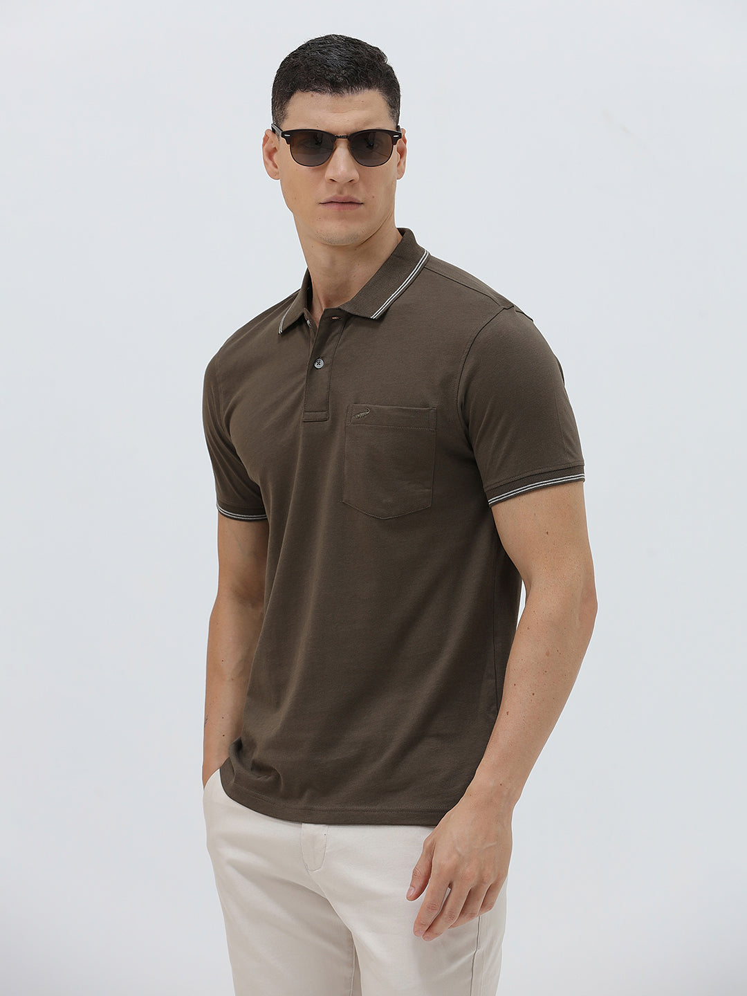 A man wearing a dark brown short-sleeve polo shirt with contrasting light gray trim on the collar and sleeve cuffs, paired with off-white trousers, against a plain light gray background.