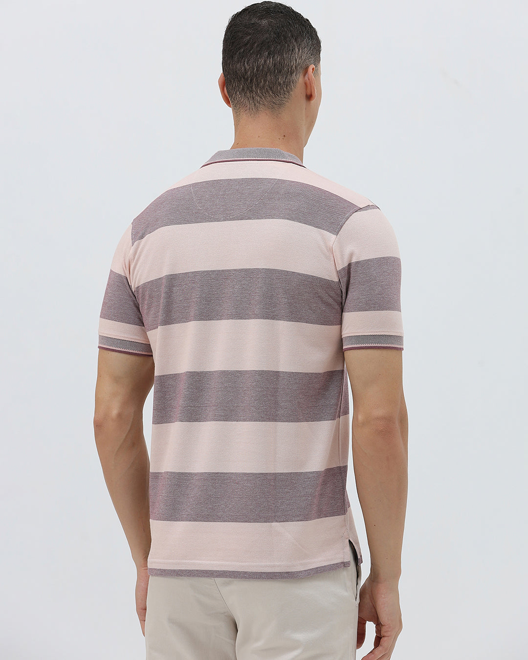 A man wearing a short-sleeve polo shirt with horizontal stripes in dusty pink and gray, paired with light beige trousers, against a plain light gray background.