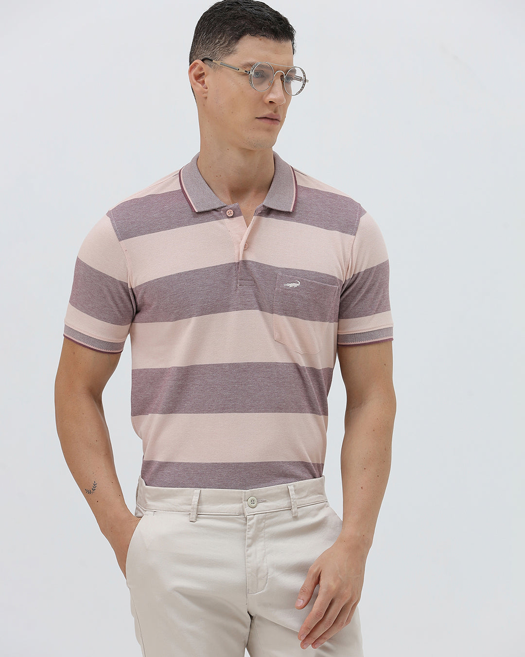 A man wearing a short-sleeve polo shirt with horizontal stripes in dusty pink and gray, paired with light beige trousers, against a plain light gray background.