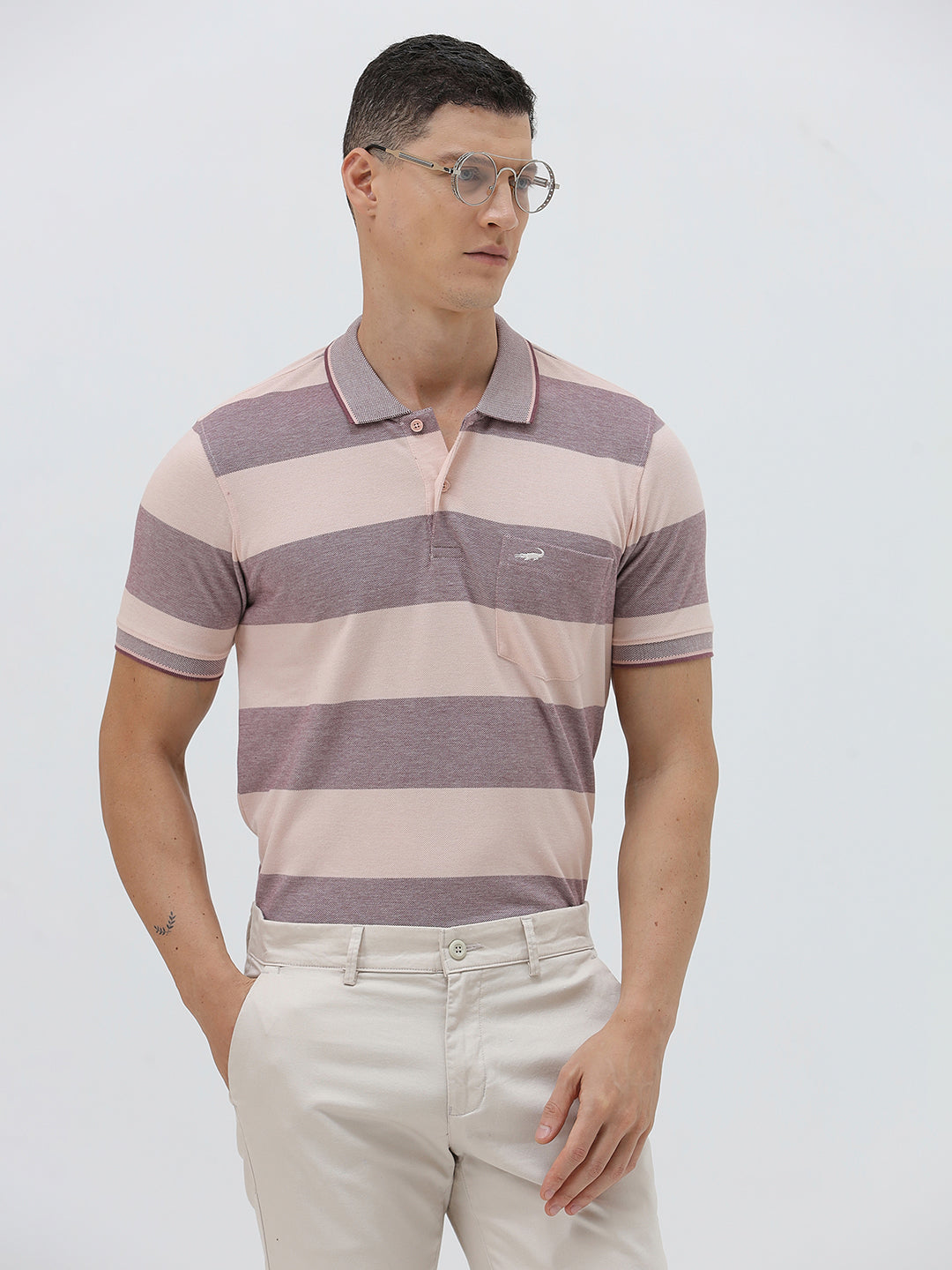 A man wearing a short-sleeve polo shirt with horizontal stripes in dusty pink and gray, paired with light beige trousers, against a plain light gray background.