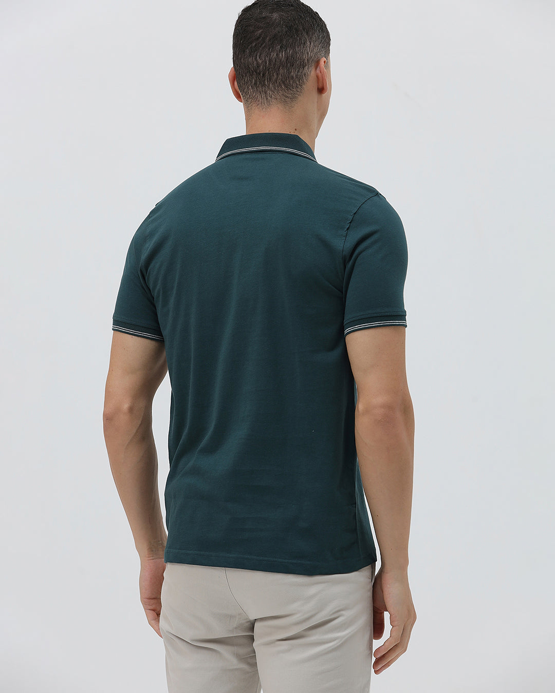 A man wearing a dark teal-green short-sleeve polo shirt with contrasting light gray piping on the collar and sleeve cuffs, paired with light beige trousers, against a plain light gray studio background.