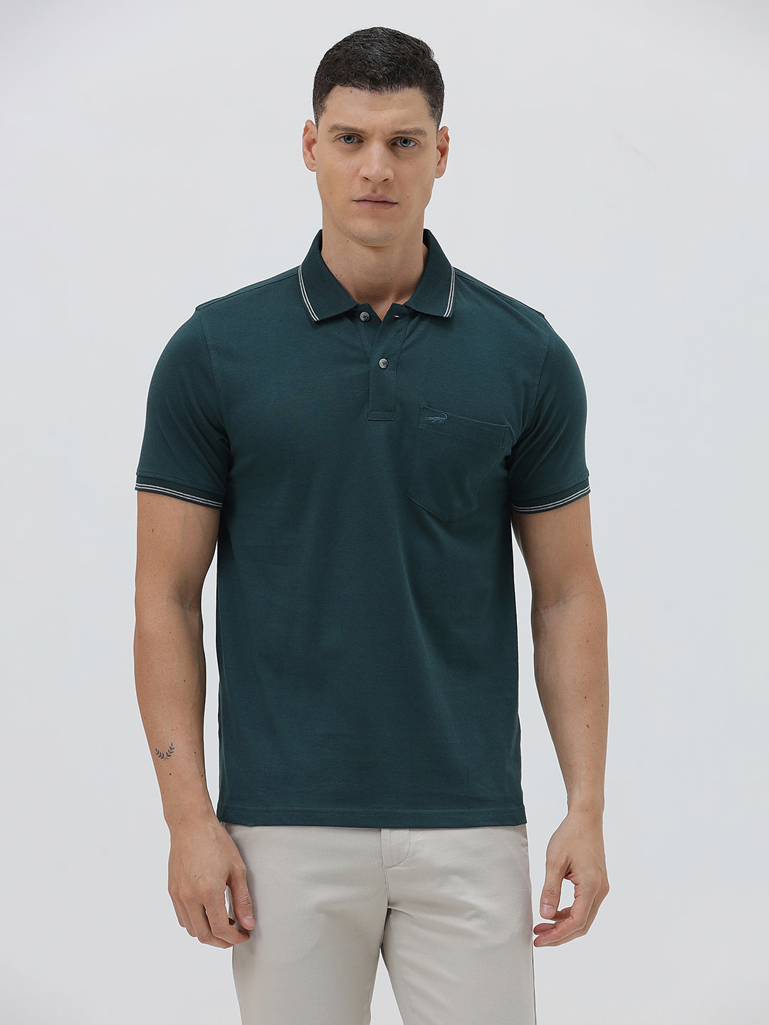 A man wearing a dark teal-green short-sleeve polo shirt with contrasting light gray piping on the collar and sleeve cuffs, paired with light beige trousers, against a plain light gray studio background.