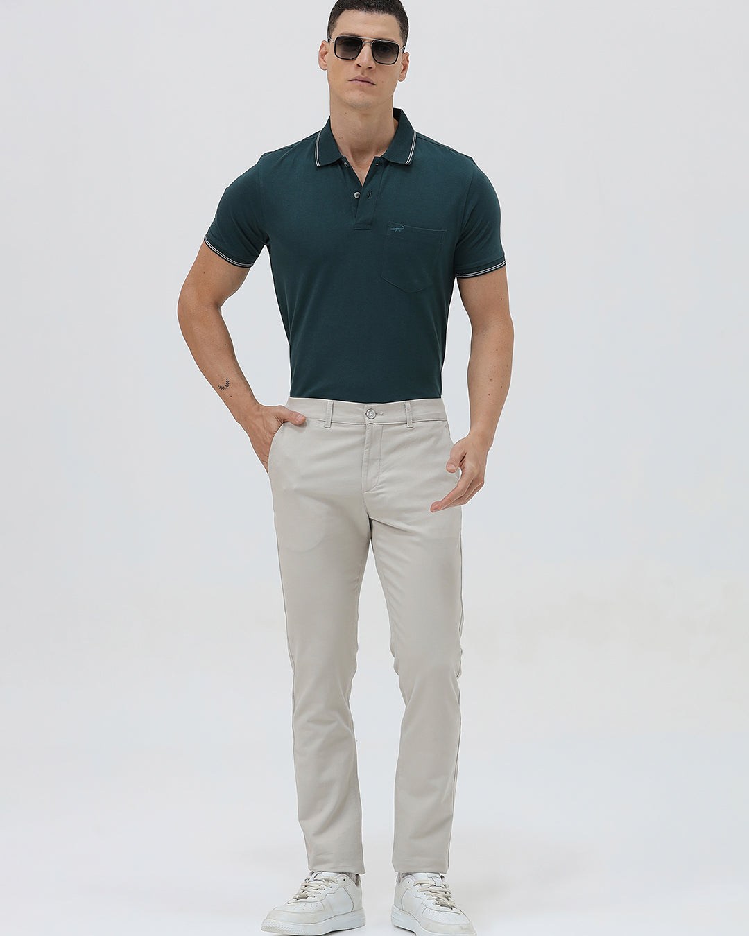 A person wearing slim-fit light beige chinos with a dark green t-shirt and white sneakers, standing against a plain white studio background.
