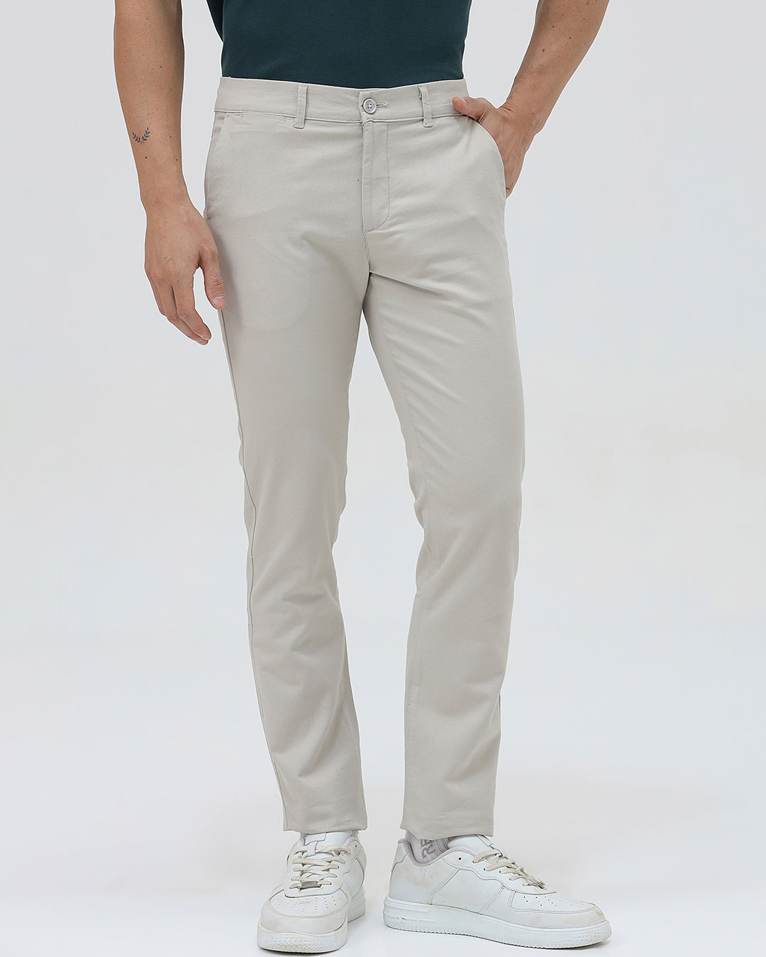 A person wearing slim-fit light beige chinos with a dark green t-shirt and white sneakers, standing against a plain white studio background.