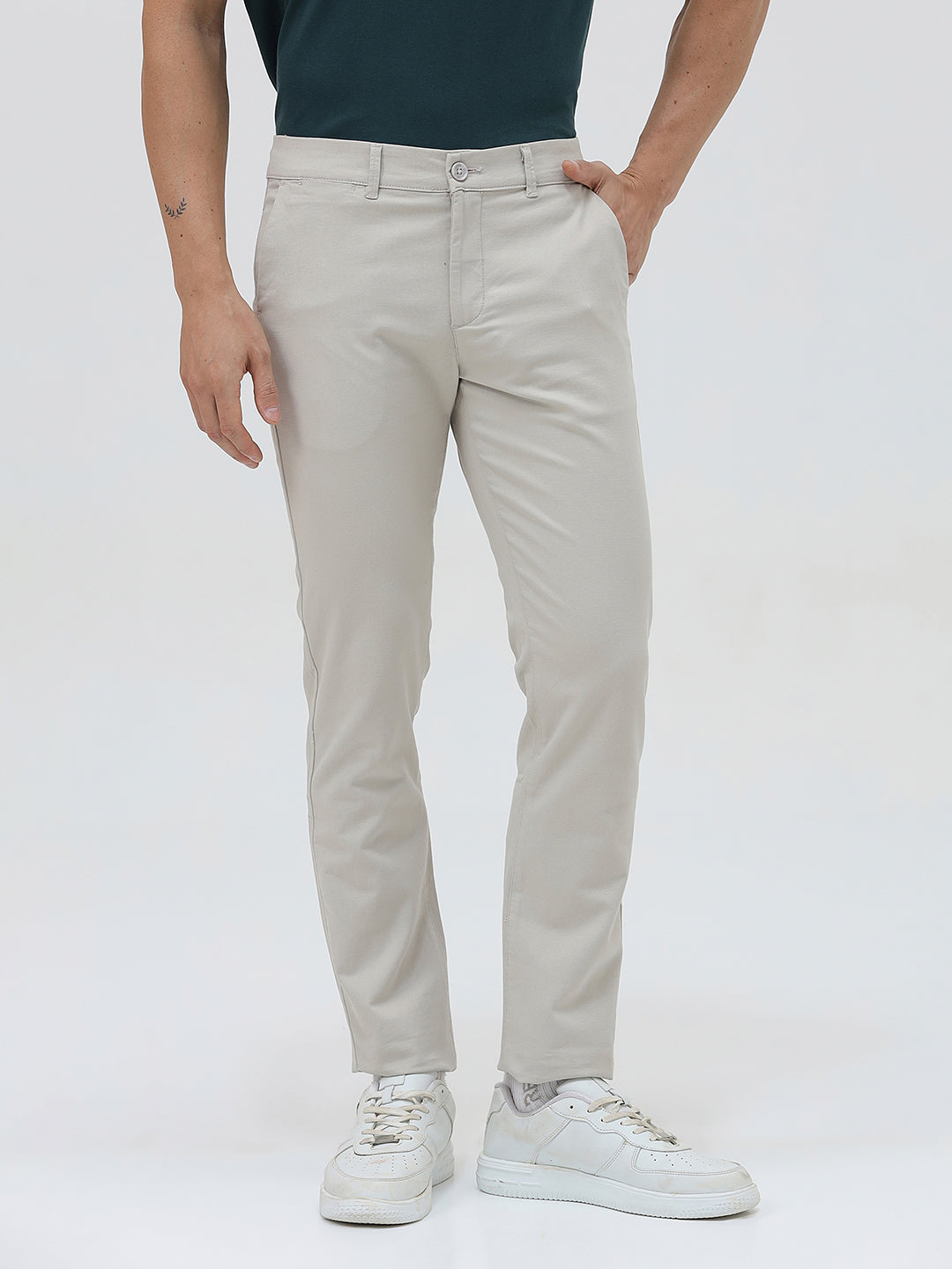 A person wearing slim-fit light beige chinos with a dark green t-shirt and white sneakers, standing against a plain white studio background.