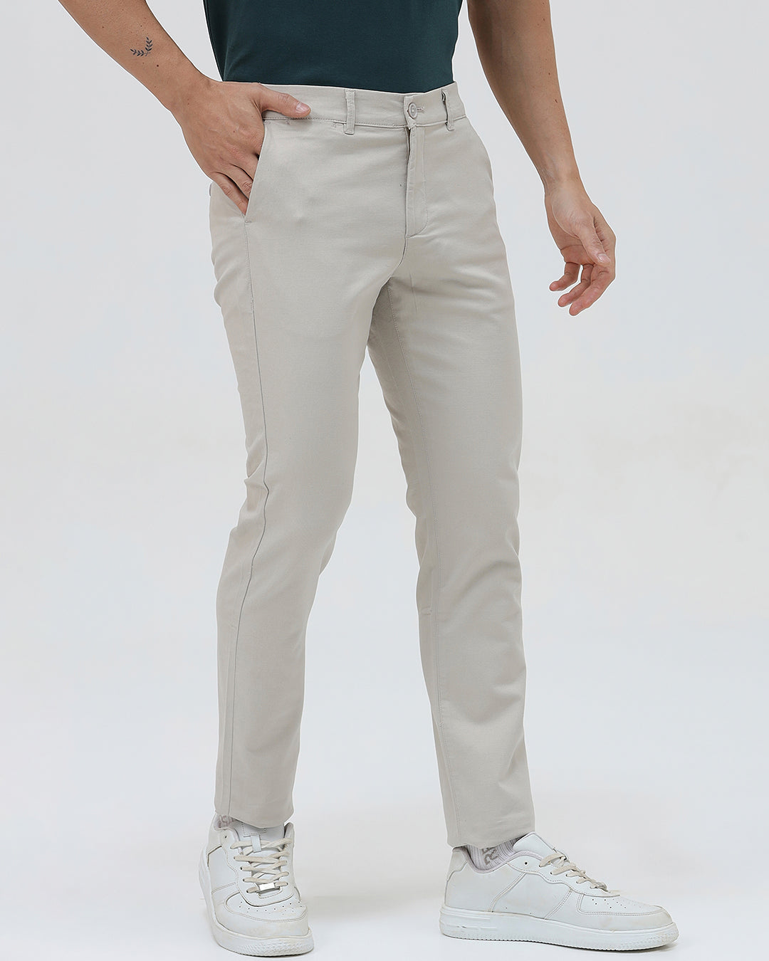 A person wearing slim-fit light beige chinos with a dark green t-shirt and white sneakers, standing against a plain white studio background.