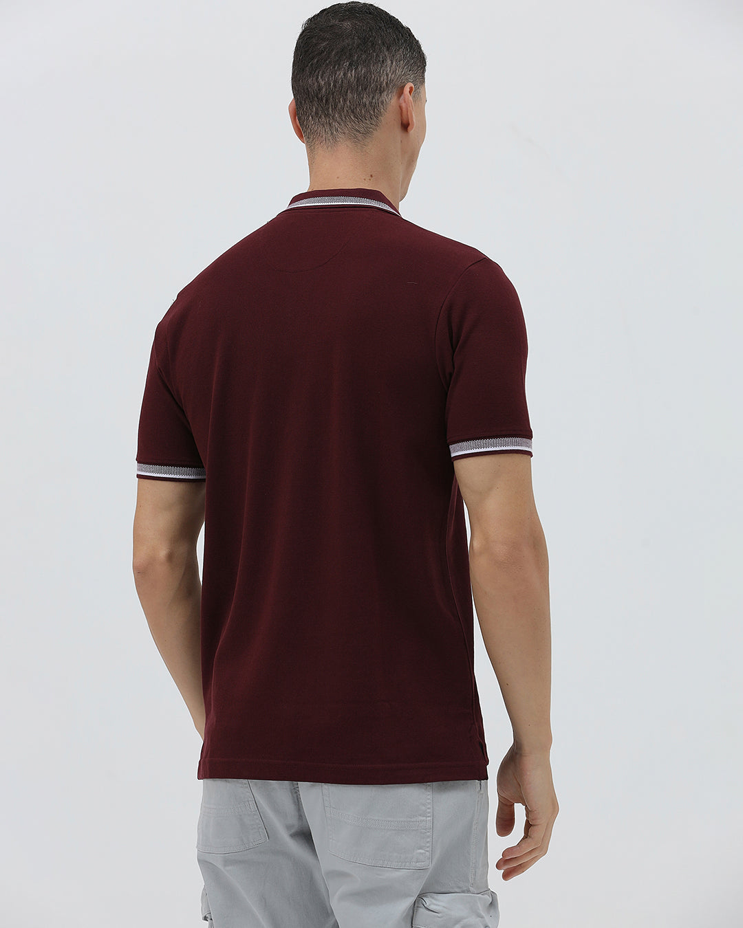 A man wearing a short-sleeve polo shirt in maroon with a contrasting light gray and white striped collar and sleeve cuffs, paired with light gray cargo pants, against a plain light gray background.