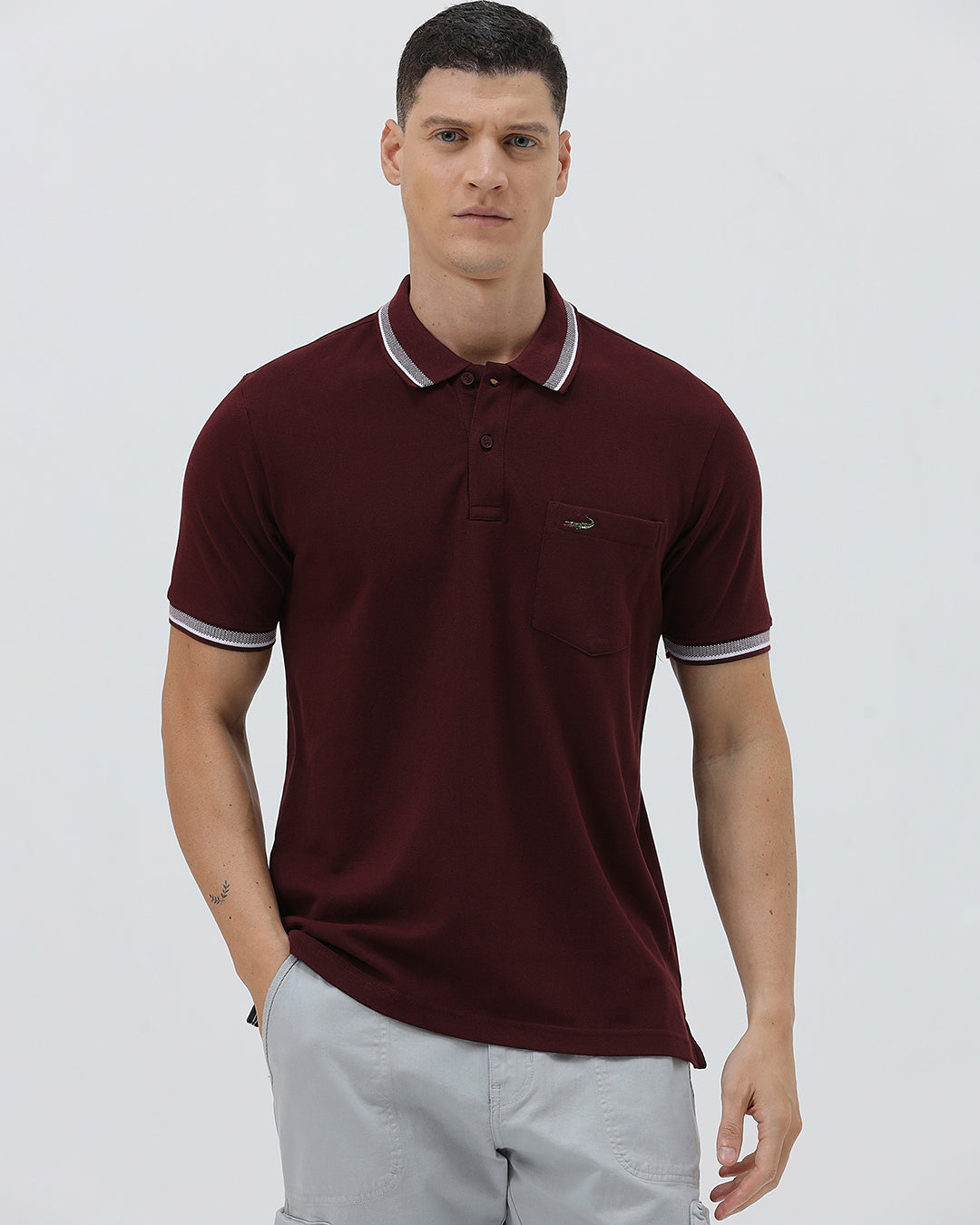 A man wearing a short-sleeve polo shirt in maroon with a contrasting light gray and white striped collar and sleeve cuffs, paired with light gray cargo pants, against a plain light gray background.