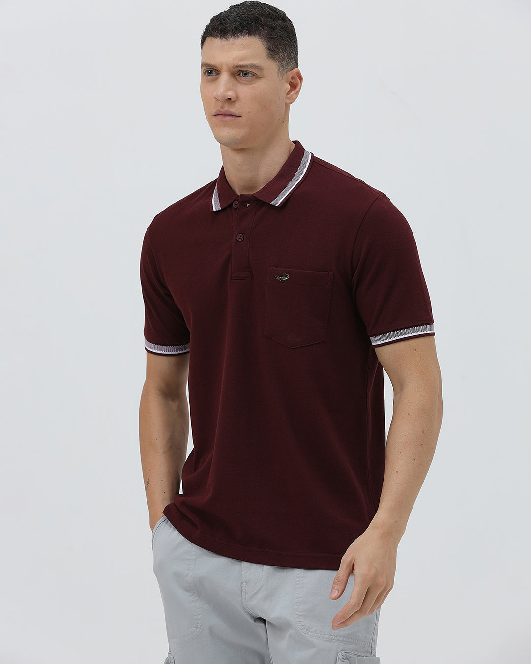 A man wearing a short-sleeve polo shirt in maroon with a contrasting light gray and white striped collar and sleeve cuffs, paired with light gray cargo pants, against a plain light gray background.