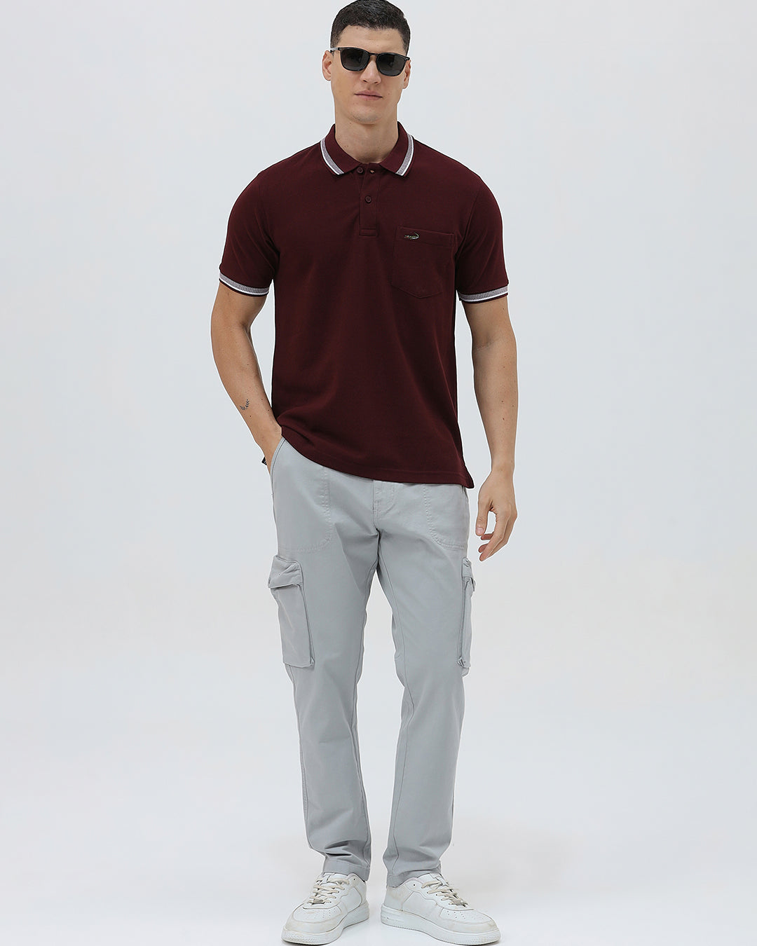 A man wearing a short-sleeve polo shirt in maroon with a contrasting light gray and white striped collar and sleeve cuffs, paired with light gray cargo pants, against a plain light gray background.