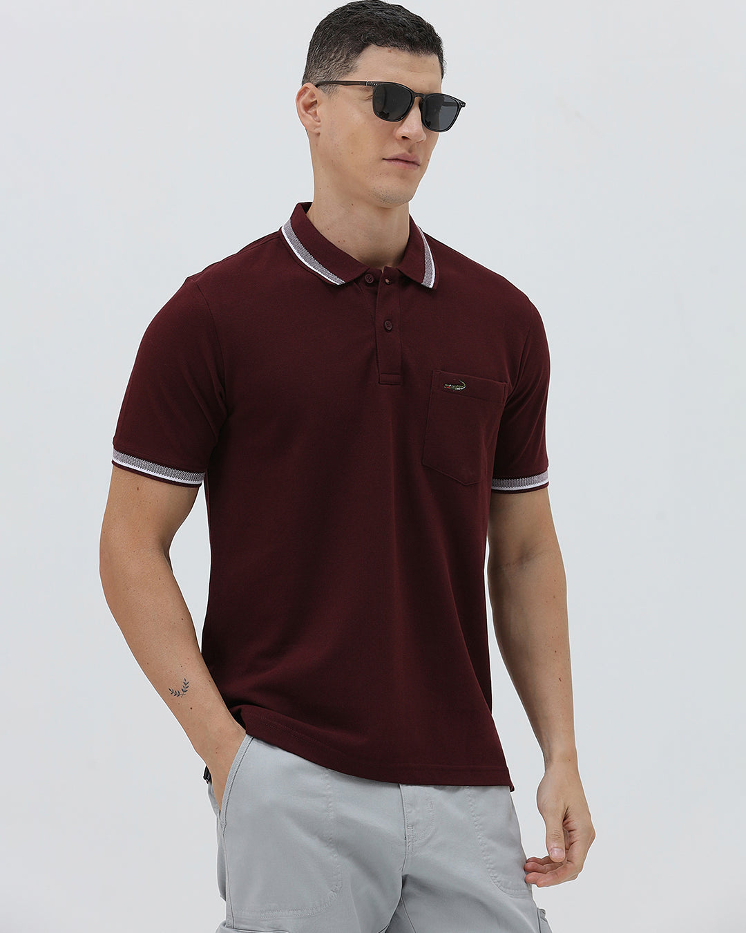 A man wearing a short-sleeve polo shirt in maroon with a contrasting light gray and white striped collar and sleeve cuffs, paired with light gray cargo pants, against a plain light gray background.