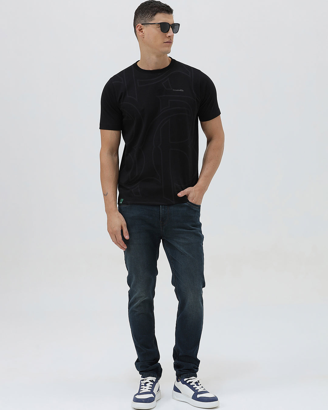 A person wearing dark blue slim-fit denim jeans, paired with a black t-shirt and navy and white sneakers, standing against a plain white studio background.