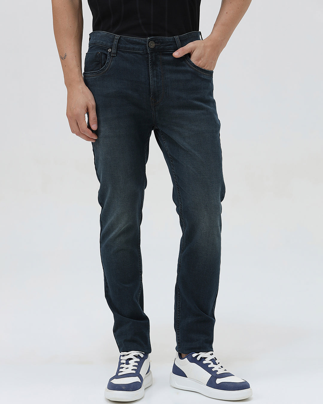 A person wearing dark blue slim-fit denim jeans, paired with a black t-shirt and navy and white sneakers, standing against a plain white studio background.
