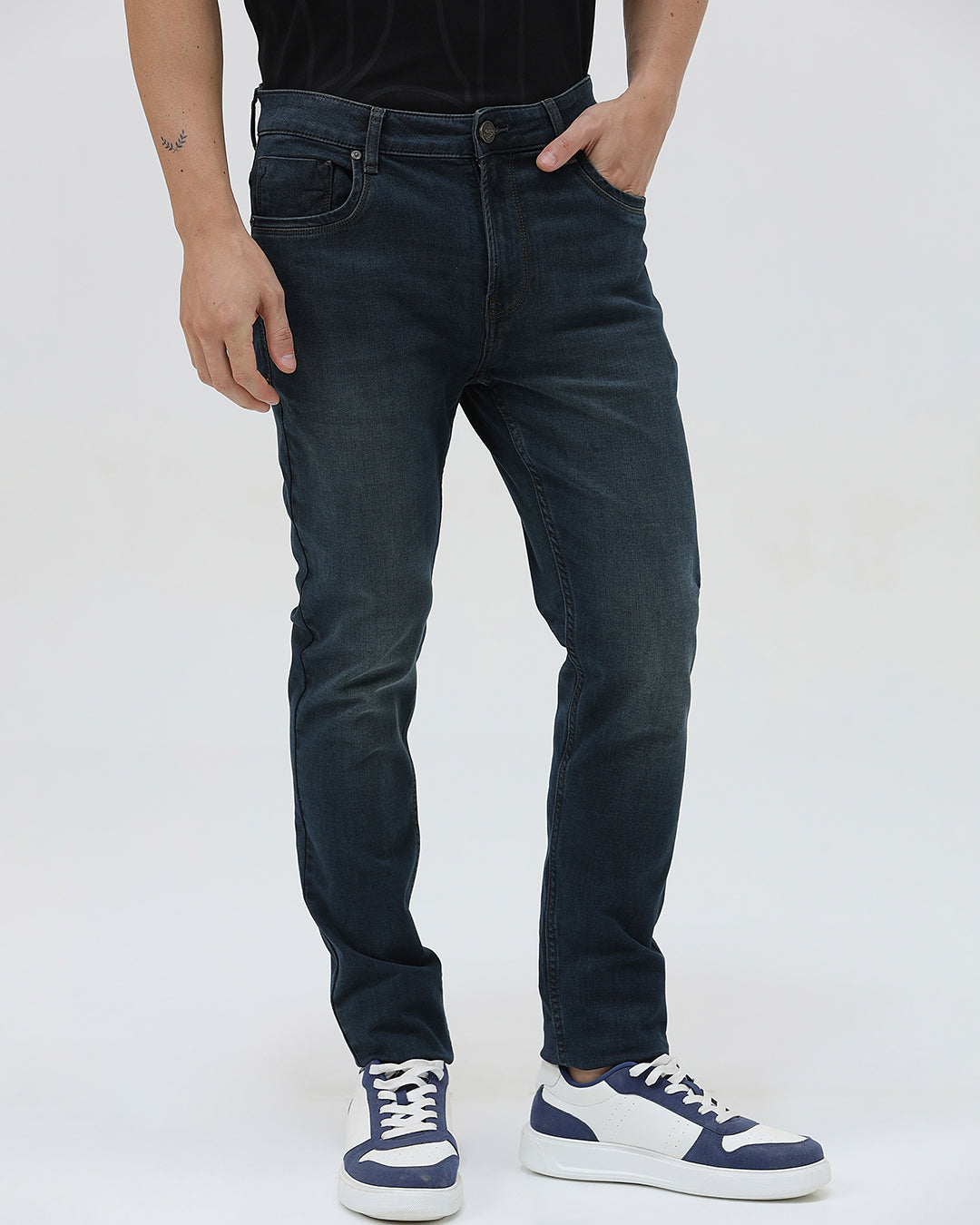 A person wearing dark blue slim-fit denim jeans, paired with a black t-shirt and navy and white sneakers, standing against a plain white studio background.