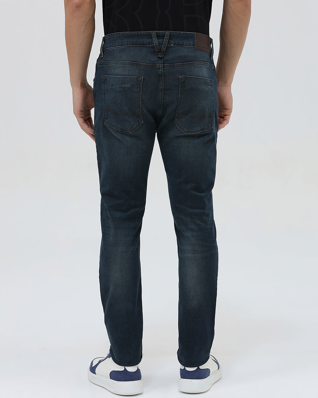 A person wearing dark blue slim-fit denim jeans, paired with a black t-shirt and navy and white sneakers, standing against a plain white studio background.