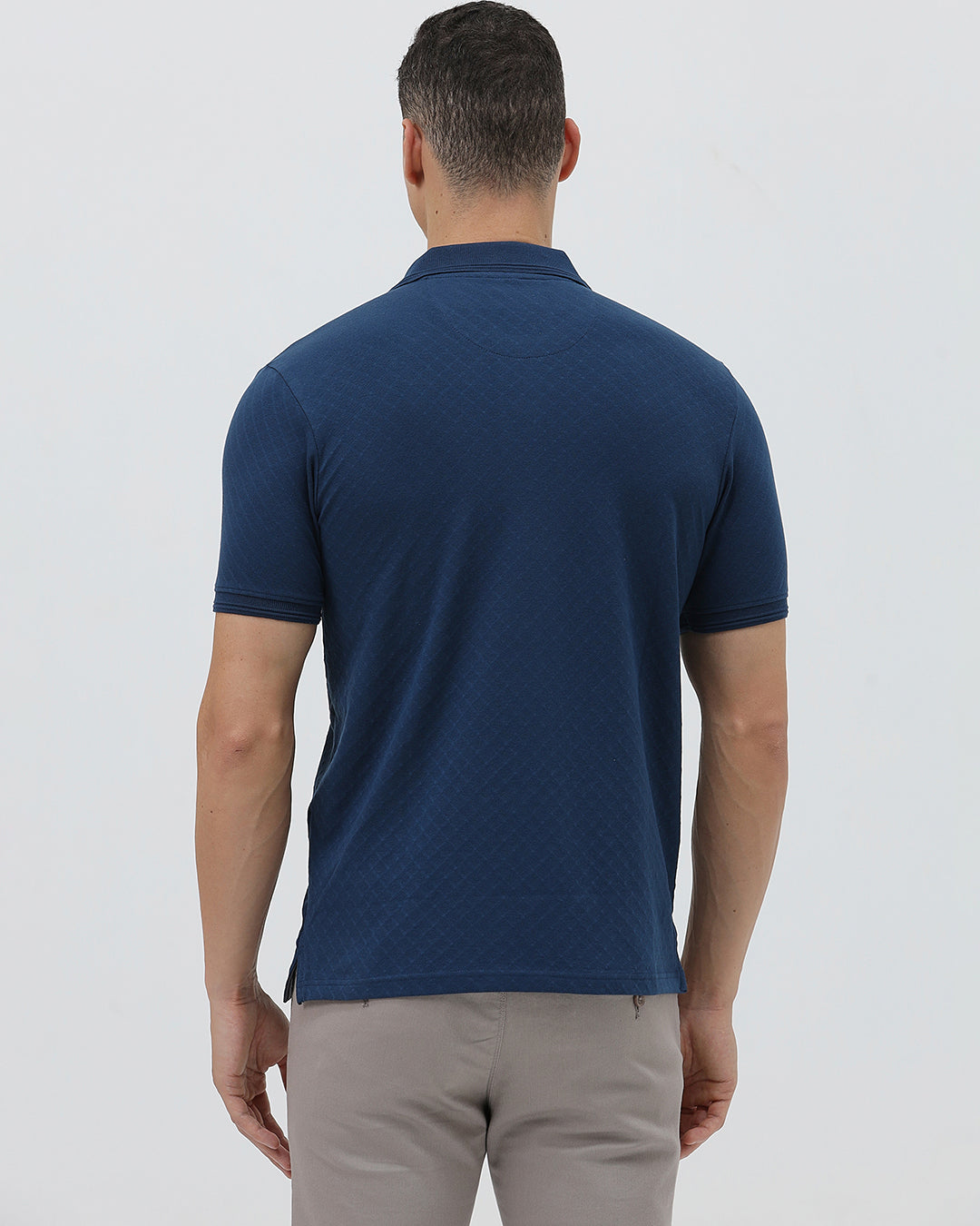A man wearing a navy blue short-sleeve polo shirt with a chest pocket, paired with light gray trousers, standing against a plain light gray studio background.