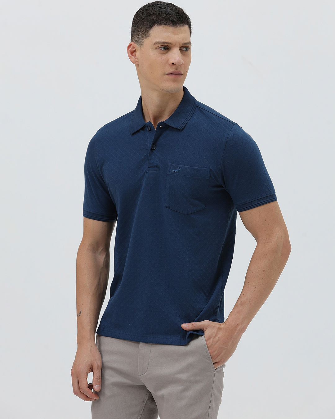 A man wearing a navy blue short-sleeve polo shirt with a chest pocket, paired with light gray trousers, standing against a plain light gray studio background.