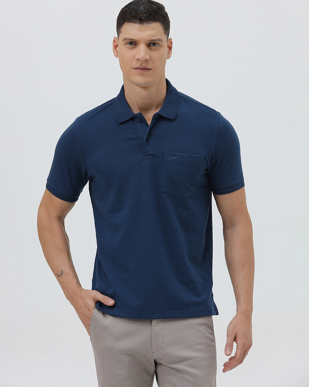 A man wearing a navy blue short-sleeve polo shirt with a chest pocket, paired with light gray trousers, standing against a plain light gray studio background.