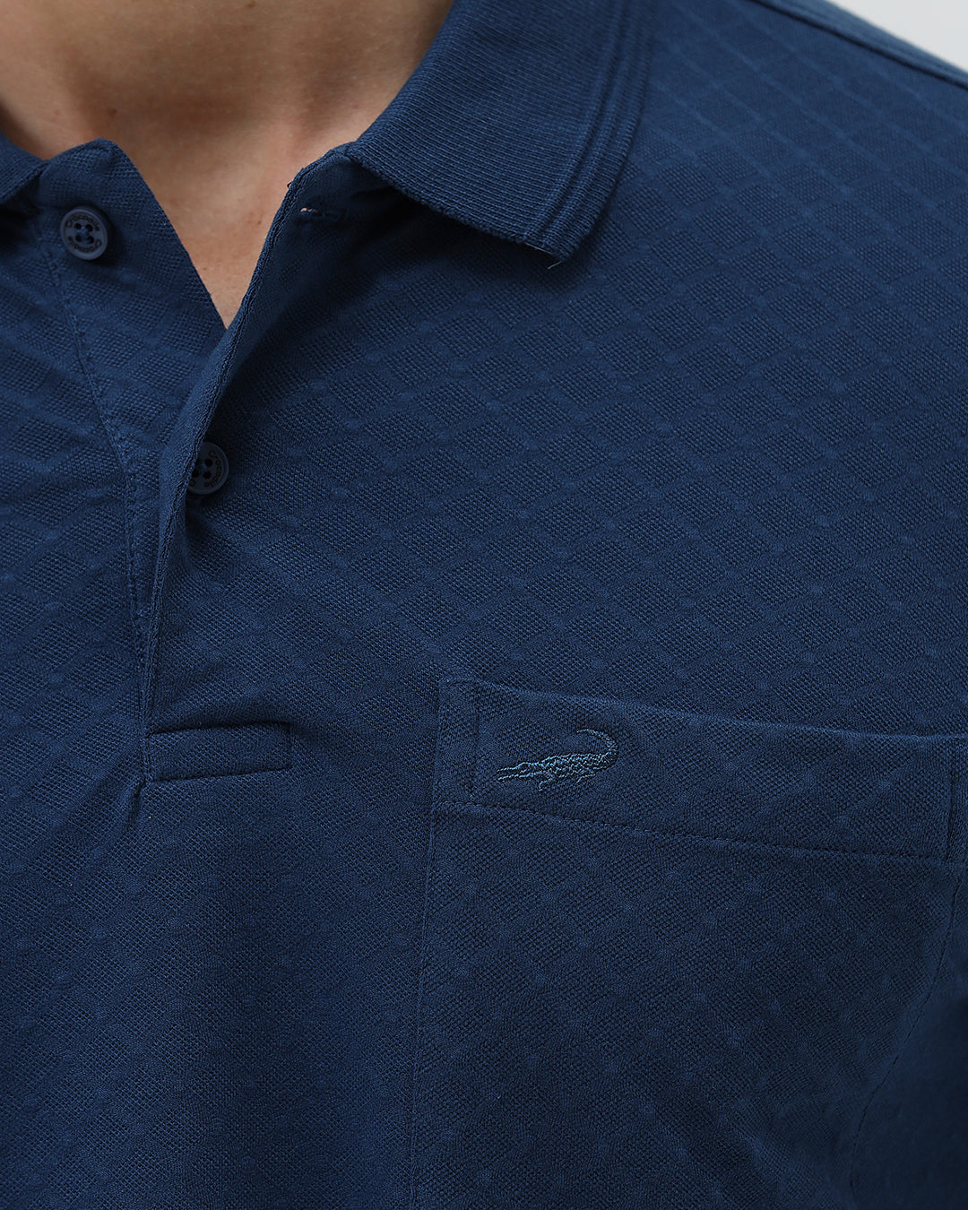 A man wearing a navy blue short-sleeve polo shirt with a chest pocket, paired with light gray trousers, standing against a plain light gray studio background.