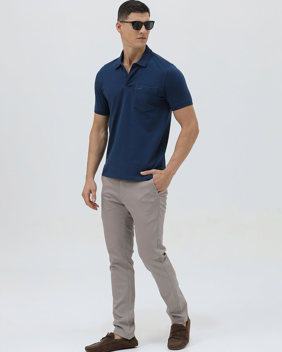 A man wearing a navy blue short-sleeve polo shirt with a chest pocket, paired with light gray trousers, standing against a plain light gray studio background.