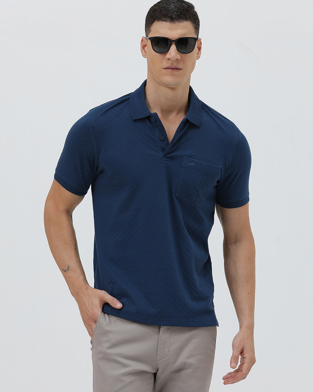 A man wearing a navy blue short-sleeve polo shirt with a chest pocket, paired with light gray trousers, standing against a plain light gray studio background.