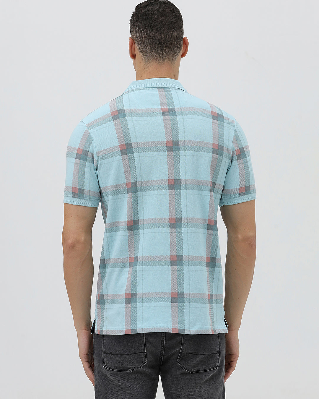 A man wearing a light blue short-sleeve polo shirt with a red and gray plaid pattern, paired with dark gray jeans, standing against a light gray studio background.