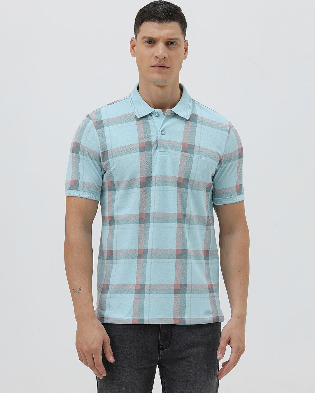 A man wearing a light blue short-sleeve polo shirt with a red and gray plaid pattern, paired with dark gray jeans, standing against a light gray studio background.