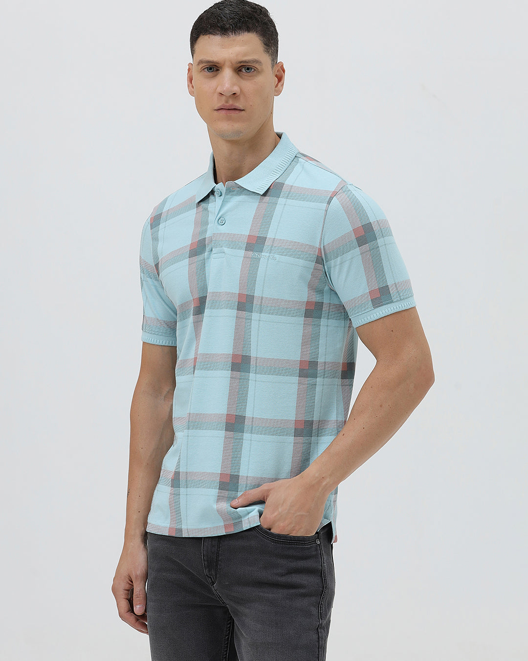A man wearing a light blue short-sleeve polo shirt with a red and gray plaid pattern, paired with dark gray jeans, standing against a light gray studio background.