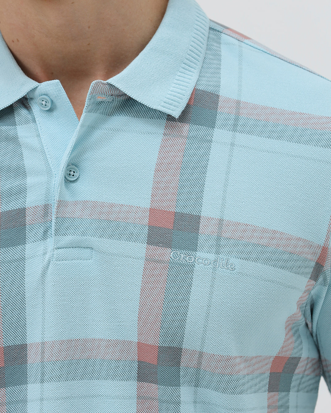 A man wearing a light blue short-sleeve polo shirt with a red and gray plaid pattern, paired with dark gray jeans, standing against a light gray studio background.