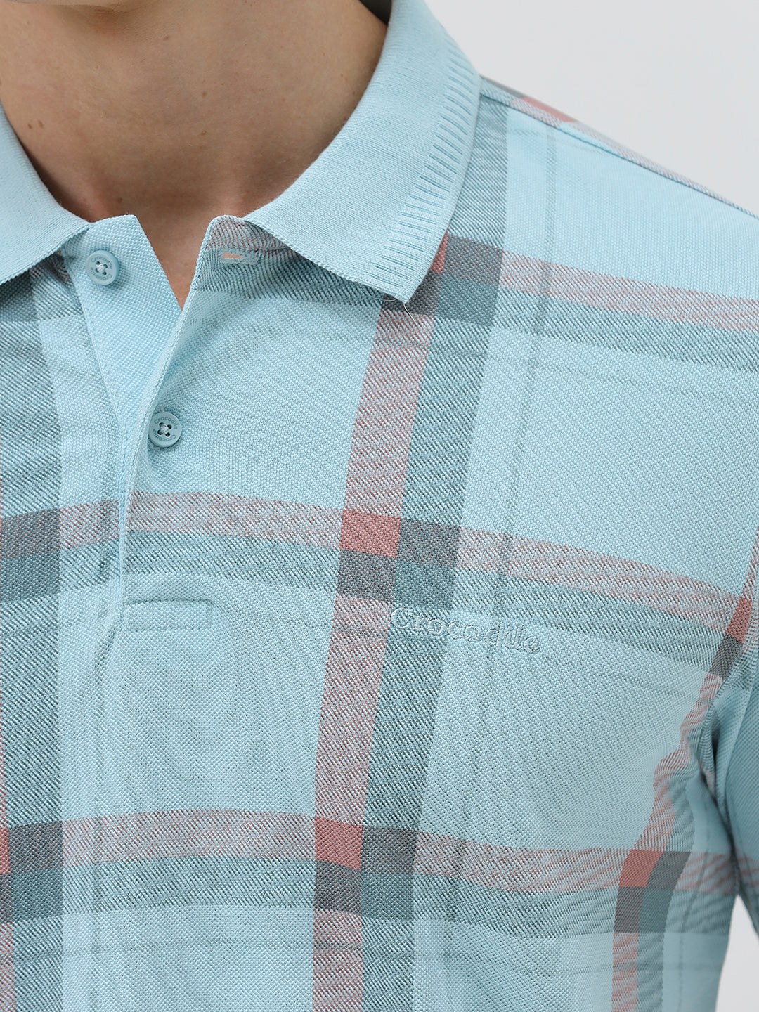 A man wearing a light blue short-sleeve polo shirt with a red and gray plaid pattern, paired with dark gray jeans, standing against a light gray studio background.