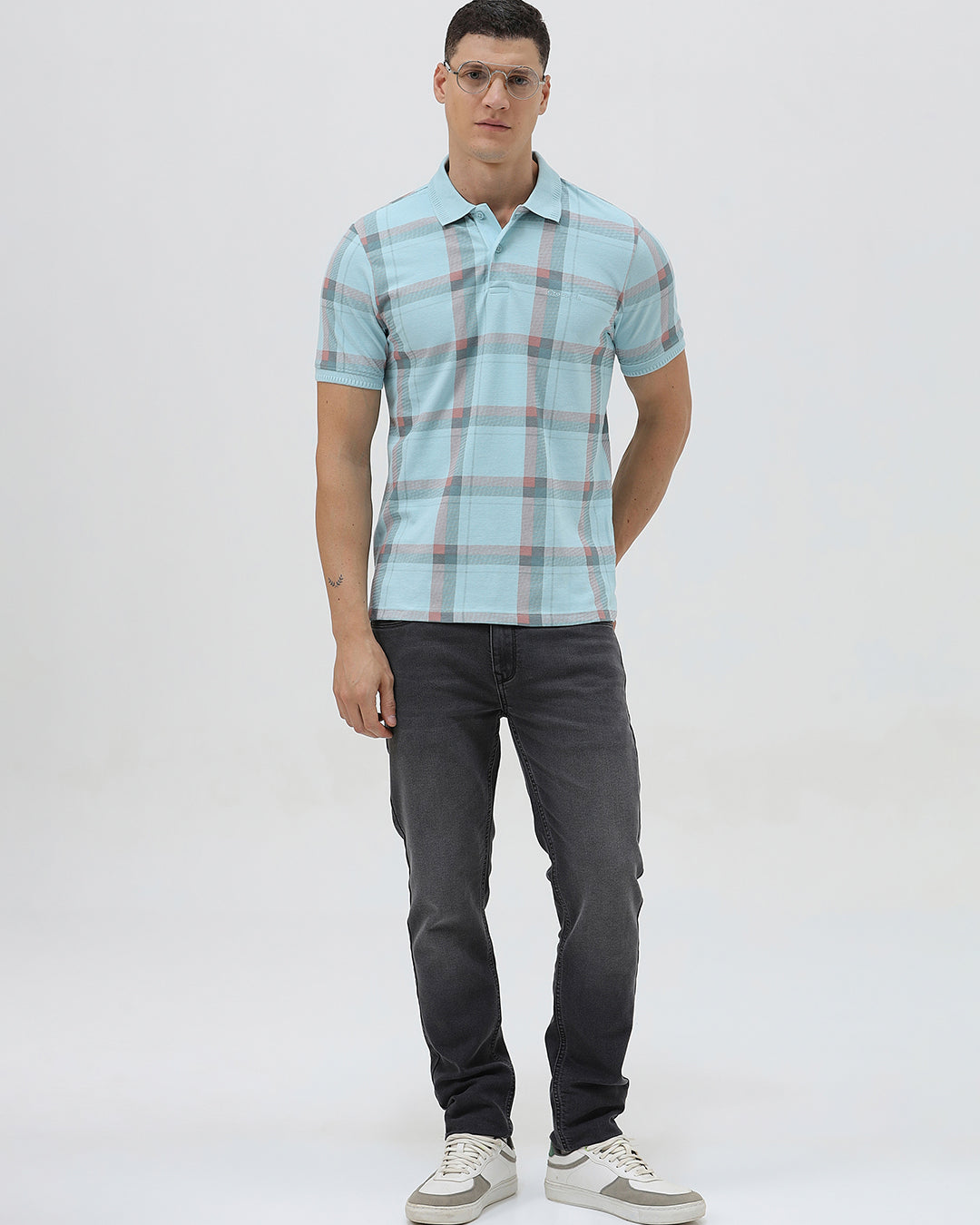 A man wearing a light blue short-sleeve polo shirt with a red and gray plaid pattern, paired with dark gray jeans, standing against a light gray studio background.