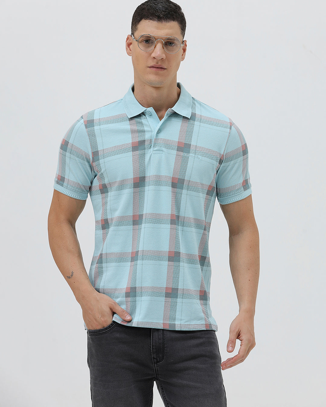 A man wearing a light blue short-sleeve polo shirt with a red and gray plaid pattern, paired with dark gray jeans, standing against a light gray studio background.