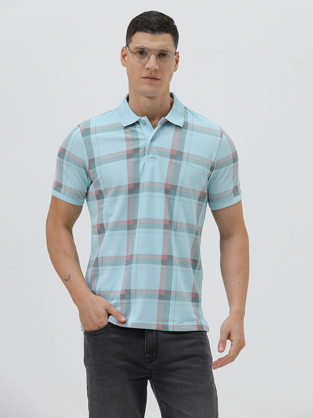 A man wearing a light blue short-sleeve polo shirt with a red and gray plaid pattern, paired with dark gray jeans, standing against a light gray studio background.