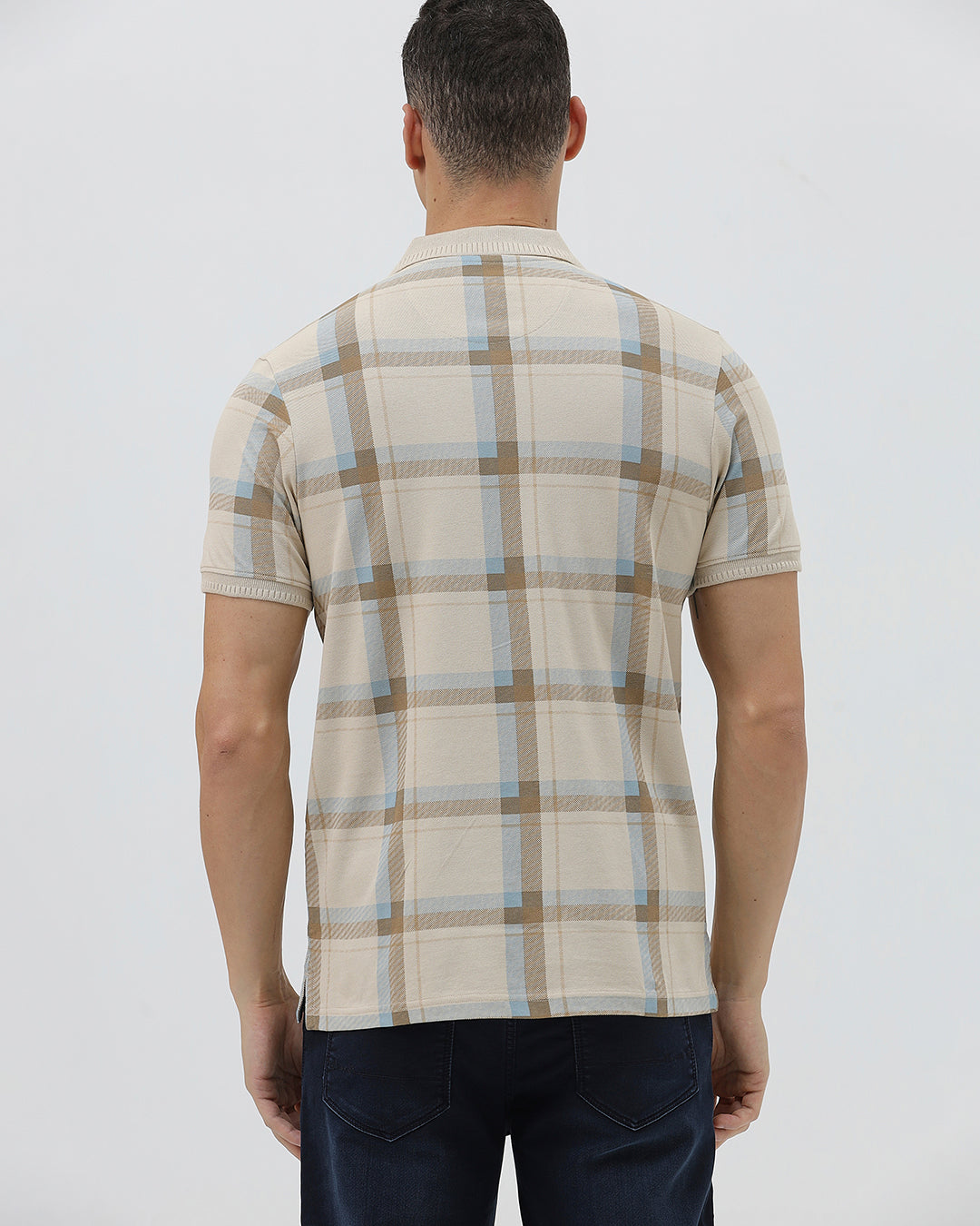 A man wearing a beige short-sleeve polo shirt with a brown and light blue plaid pattern, paired with dark blue jeans, standing against a light gray studio background.