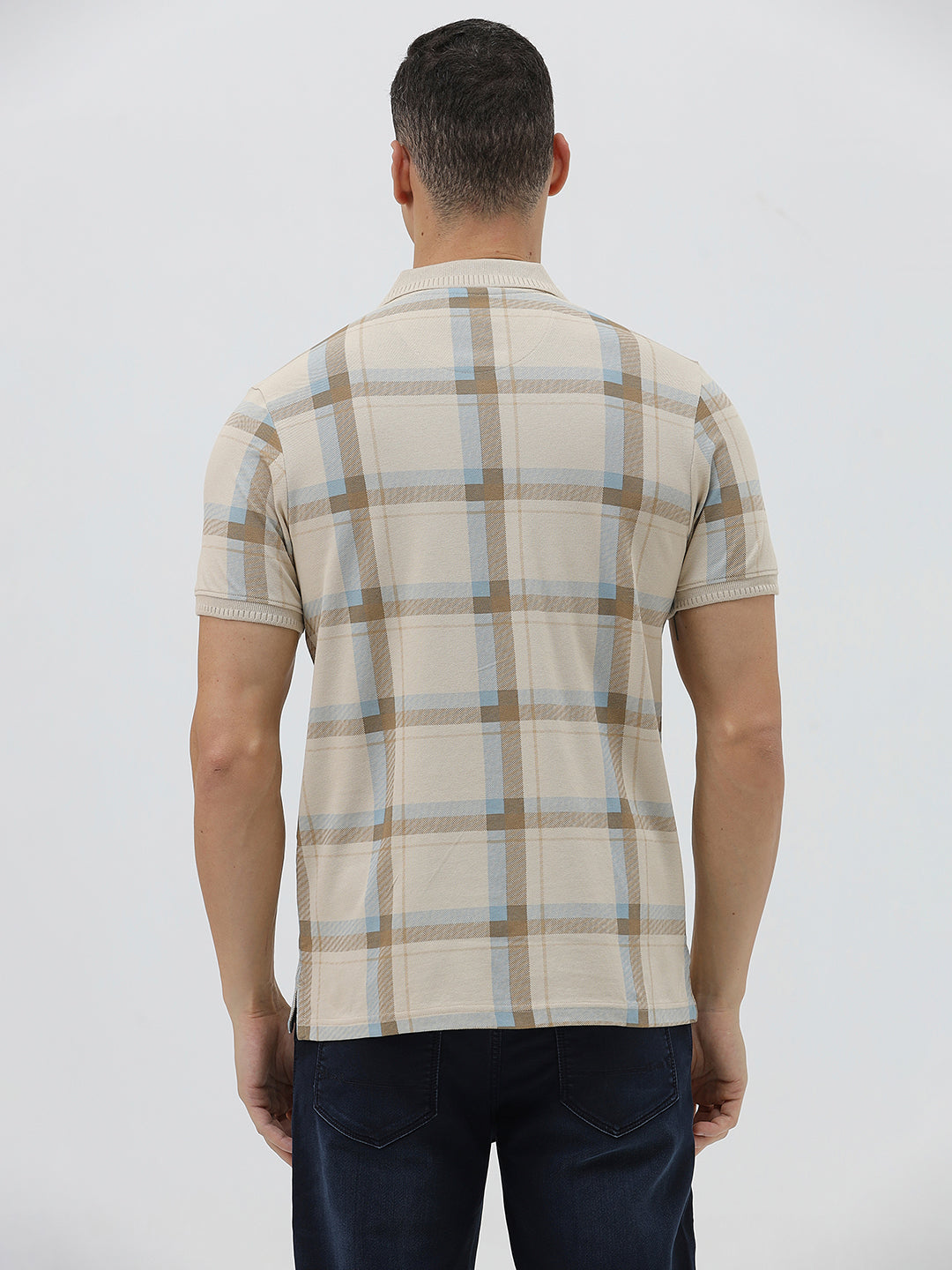 A man wearing a beige short-sleeve polo shirt with a brown and light blue plaid pattern, paired with dark blue jeans, standing against a light gray studio background.