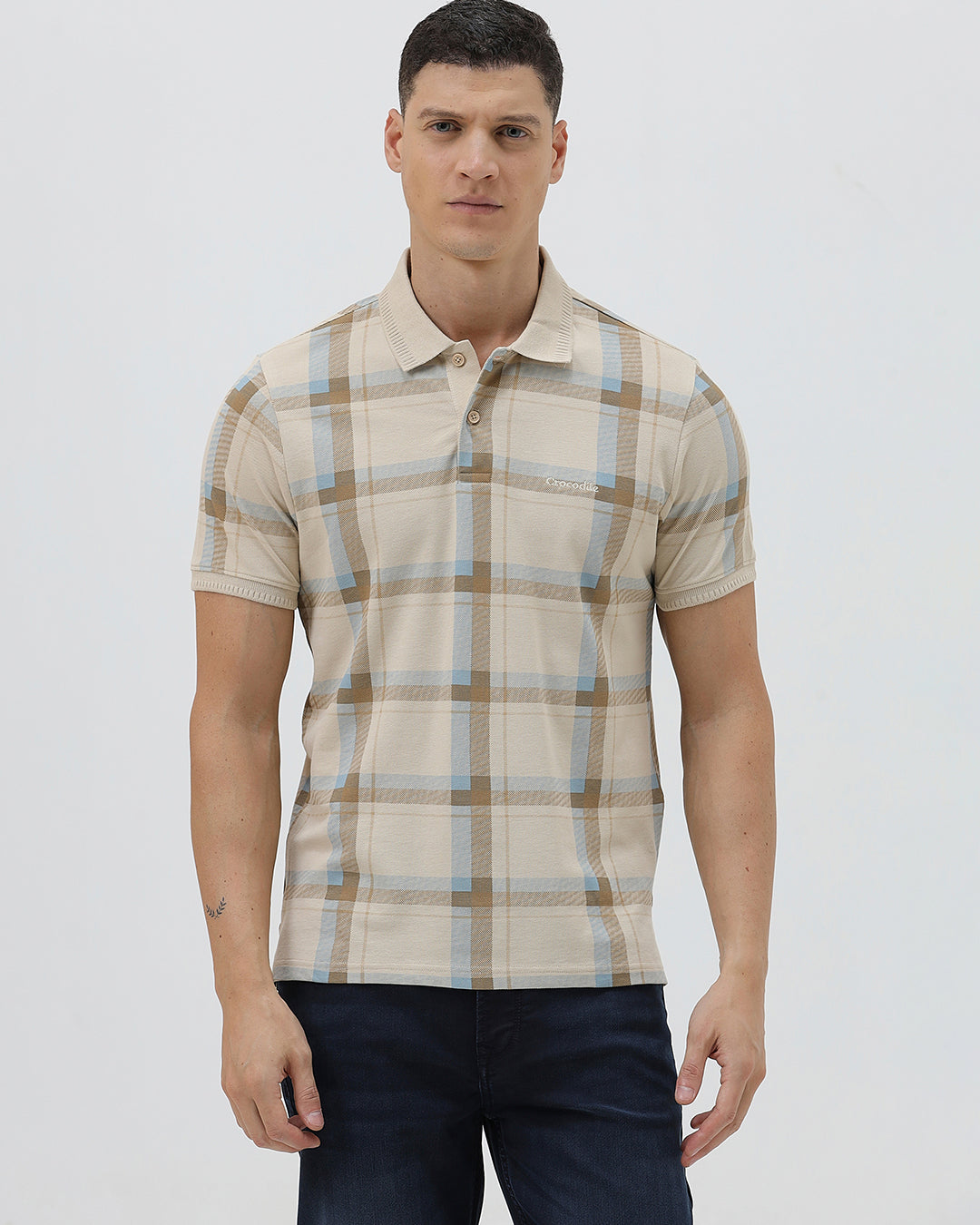 A man wearing a beige short-sleeve polo shirt with a brown and light blue plaid pattern, paired with dark blue jeans, standing against a light gray studio background.