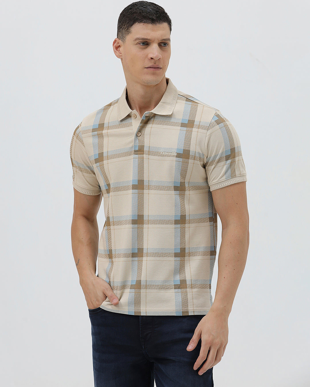 A man wearing a beige short-sleeve polo shirt with a brown and light blue plaid pattern, paired with dark blue jeans, standing against a light gray studio background.