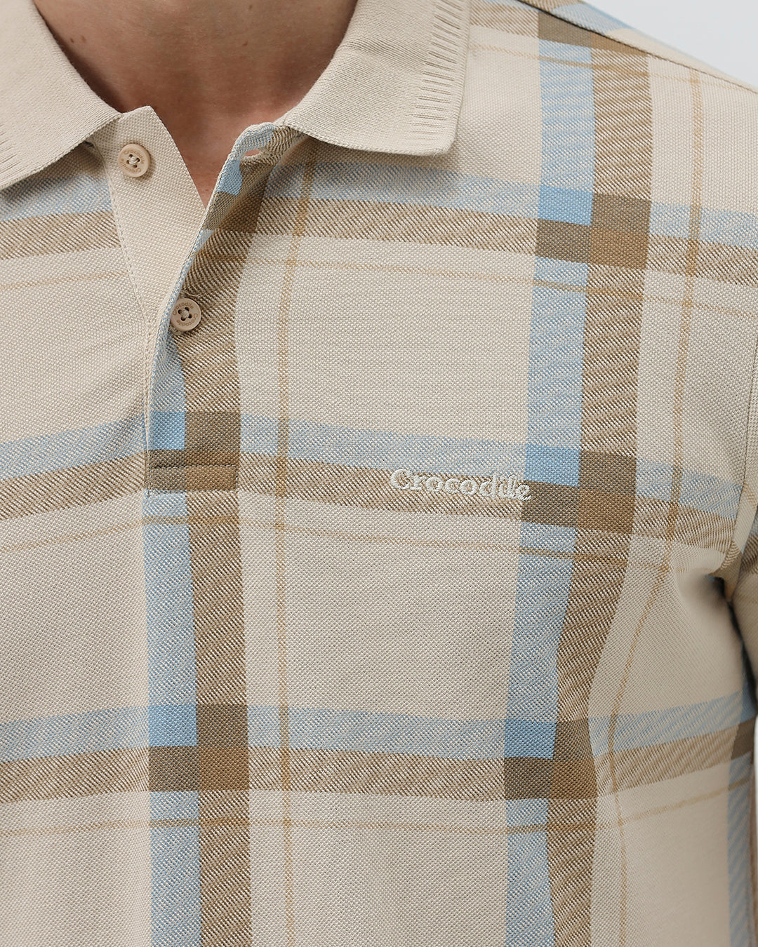 A man wearing a beige short-sleeve polo shirt with a brown and light blue plaid pattern, paired with dark blue jeans, standing against a light gray studio background.