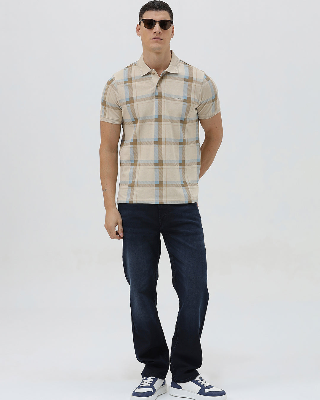 A man wearing a beige short-sleeve polo shirt with a brown and light blue plaid pattern, paired with dark blue jeans, standing against a light gray studio background.