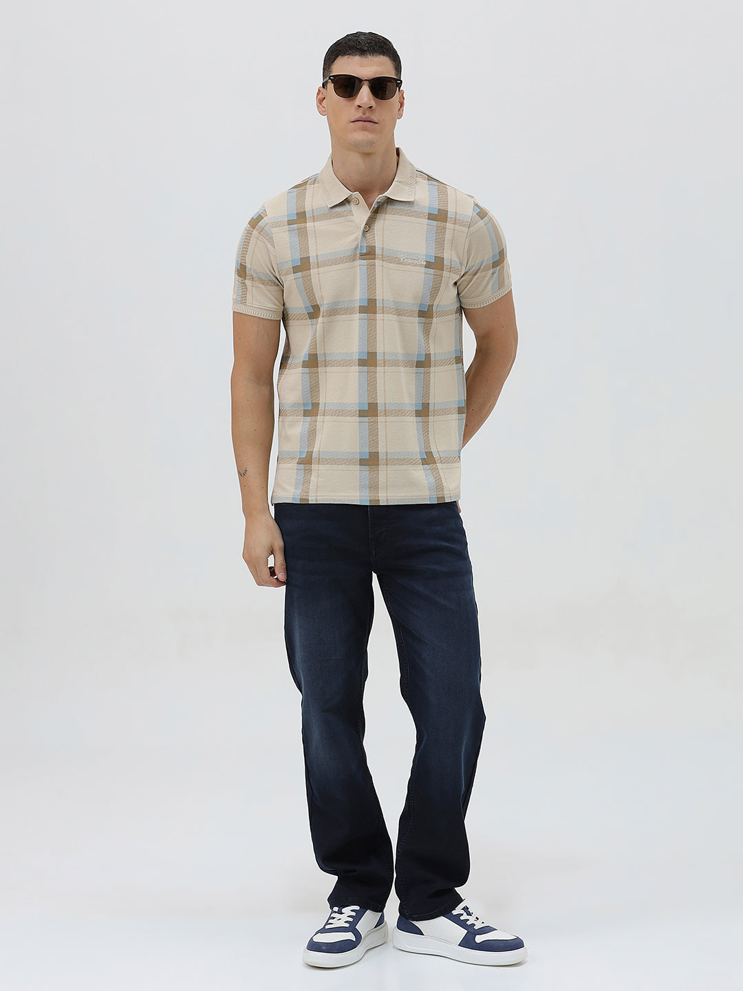 A man wearing a beige short-sleeve polo shirt with a brown and light blue plaid pattern, paired with dark blue jeans, standing against a light gray studio background.