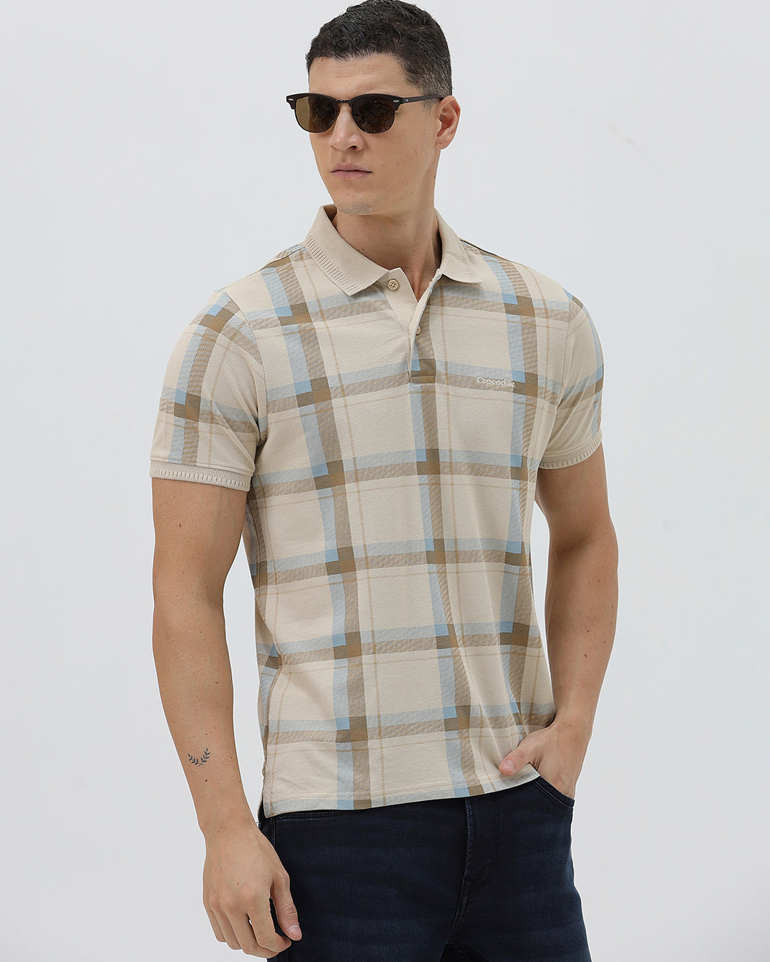 A man wearing a beige short-sleeve polo shirt with a brown and light blue plaid pattern, paired with dark blue jeans, standing against a light gray studio background.