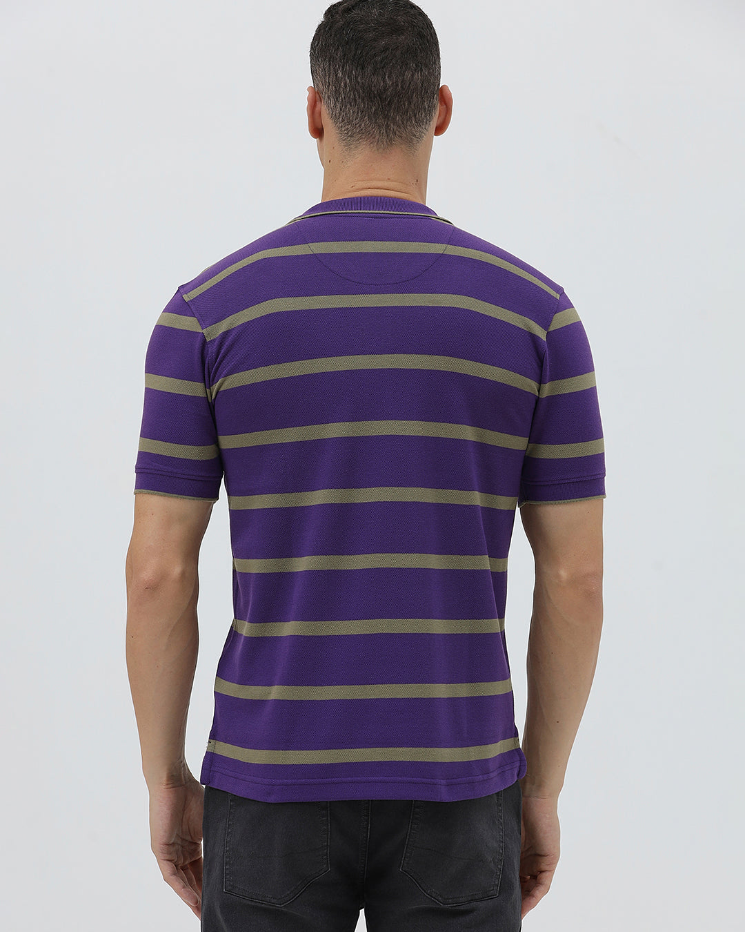A man wearing a purple short-sleeve polo shirt with horizontal olive green stripes, paired with dark gray pants, standing against a light gray studio background.