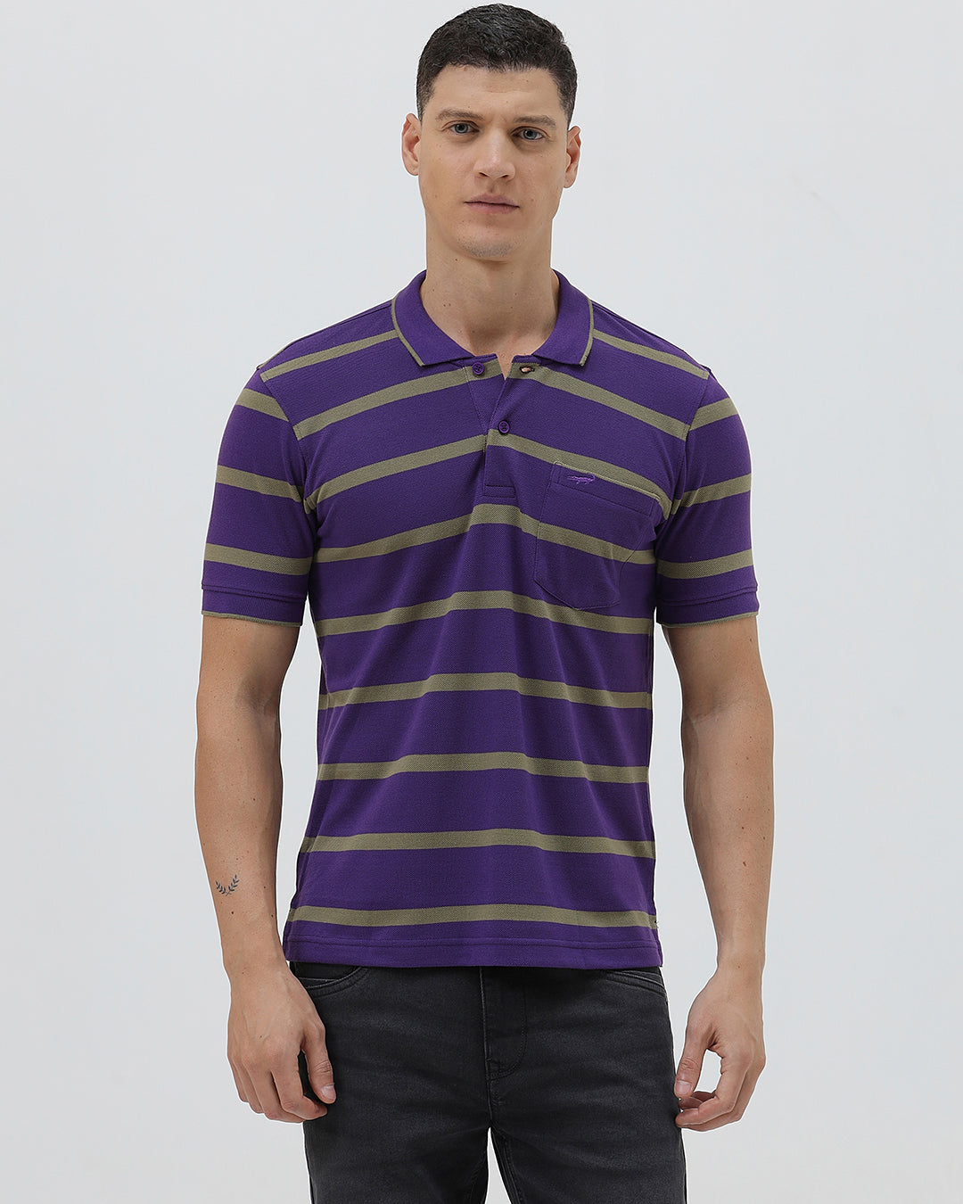 A man wearing a purple short-sleeve polo shirt with horizontal olive green stripes, paired with dark gray pants, standing against a light gray studio background.