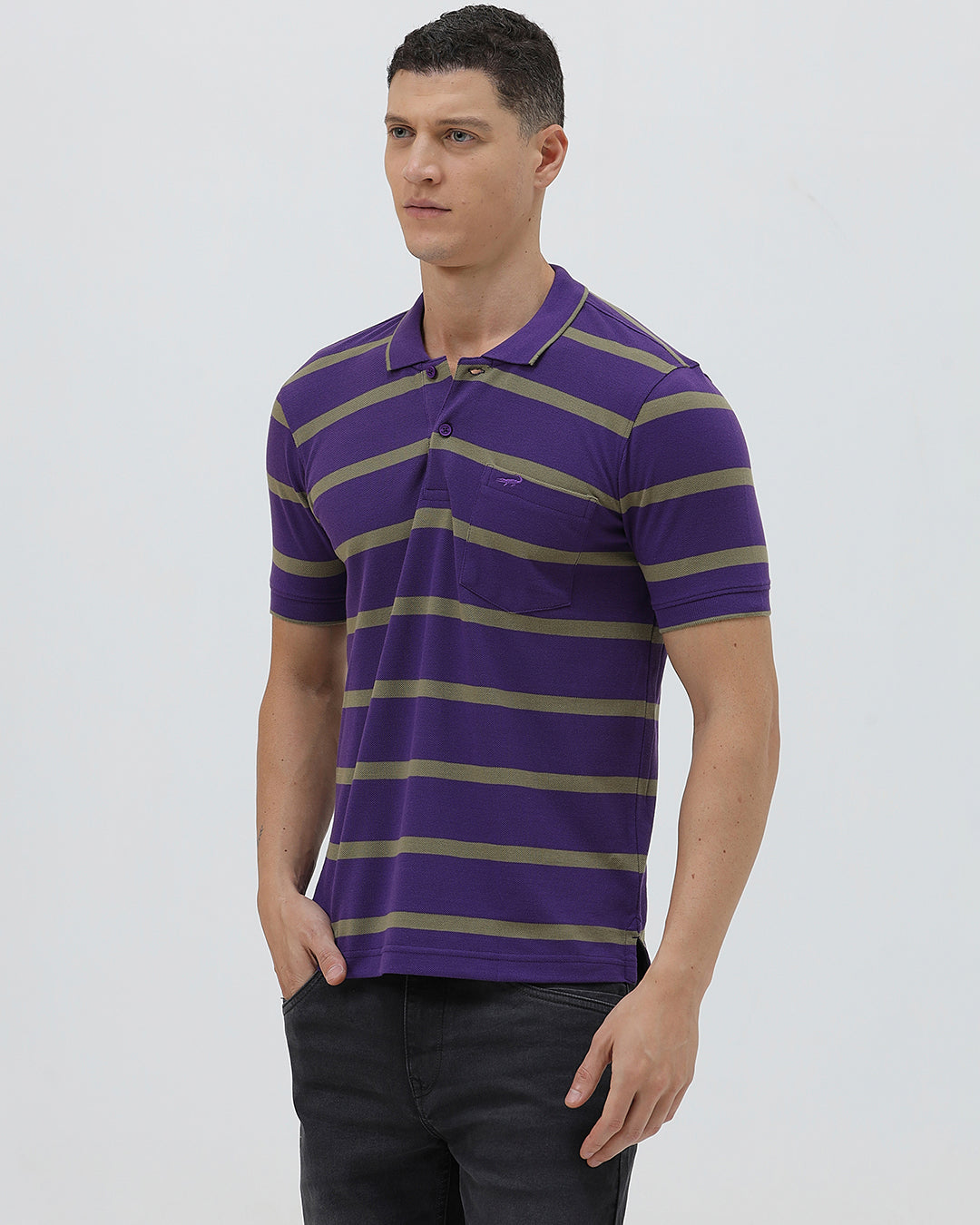 A man wearing a purple short-sleeve polo shirt with horizontal olive green stripes, paired with dark gray pants, standing against a light gray studio background.
