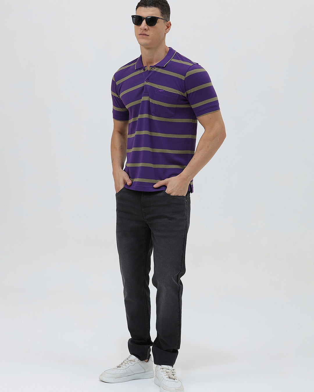 A man wearing a purple short-sleeve polo shirt with horizontal olive green stripes, paired with dark gray pants, standing against a light gray studio background.