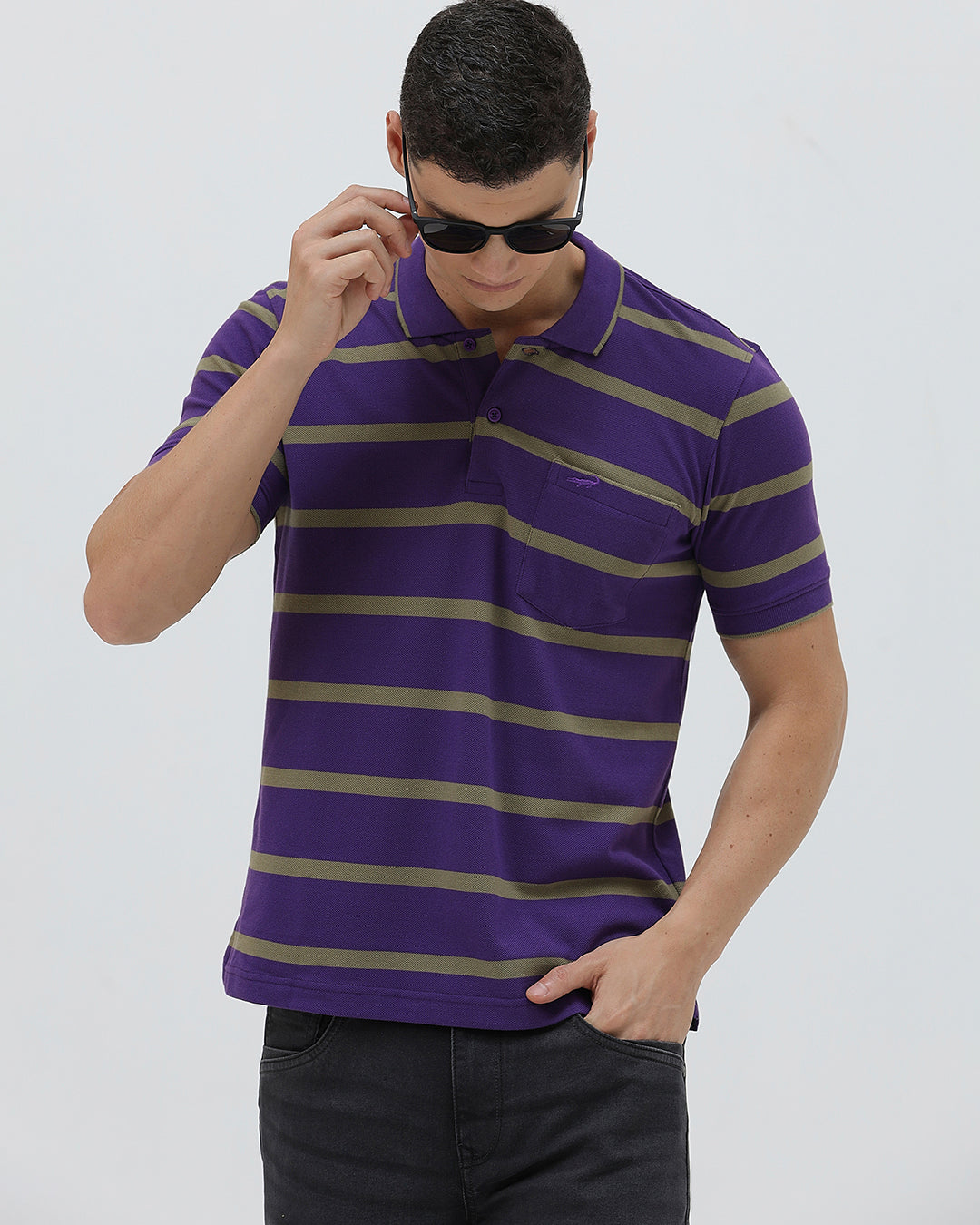 A man wearing a purple short-sleeve polo shirt with horizontal olive green stripes, paired with dark gray pants, standing against a light gray studio background.